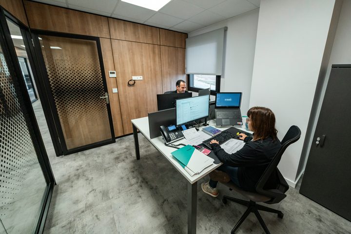 Dua staf bekerja di ruang kantor dengan layar dan dokumen di meja.
