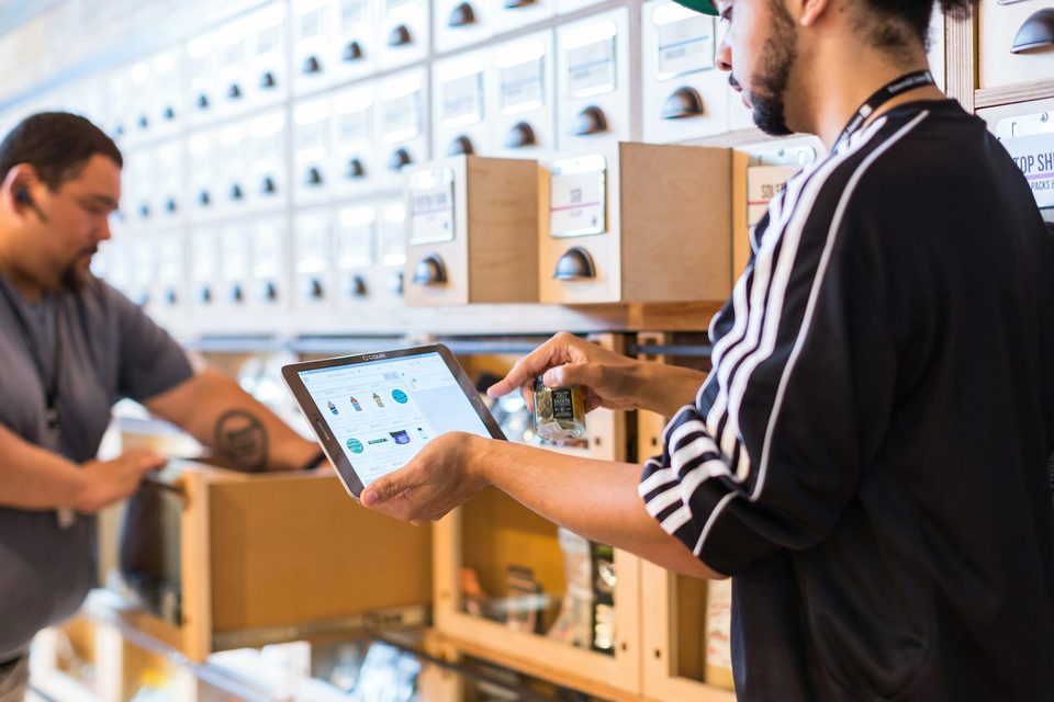 Two men in a stockroom checking a tablet next to shelves of boxes.