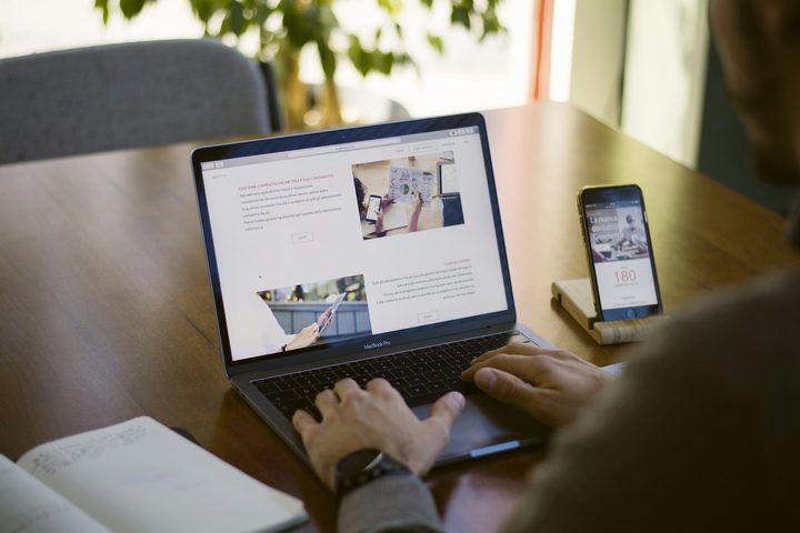 Seseorang mengetik pada laptop yang menampilkan sebuah halaman web, di samping telepon pintar pada dudukan kayu yang menampilkan versi seluler situs yang sama.