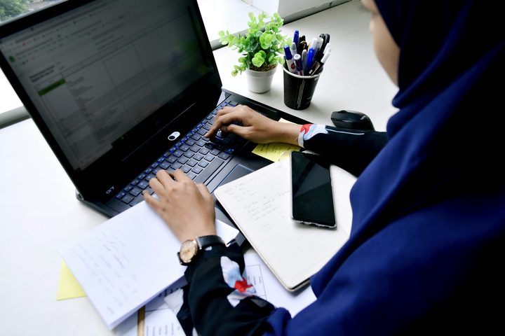 Dilihat dari atas, seorang perempuan berjilbab biru tua mengetik pada laptop di samping catatan tulisan tangan, buku catatan, dan telepon pintar.