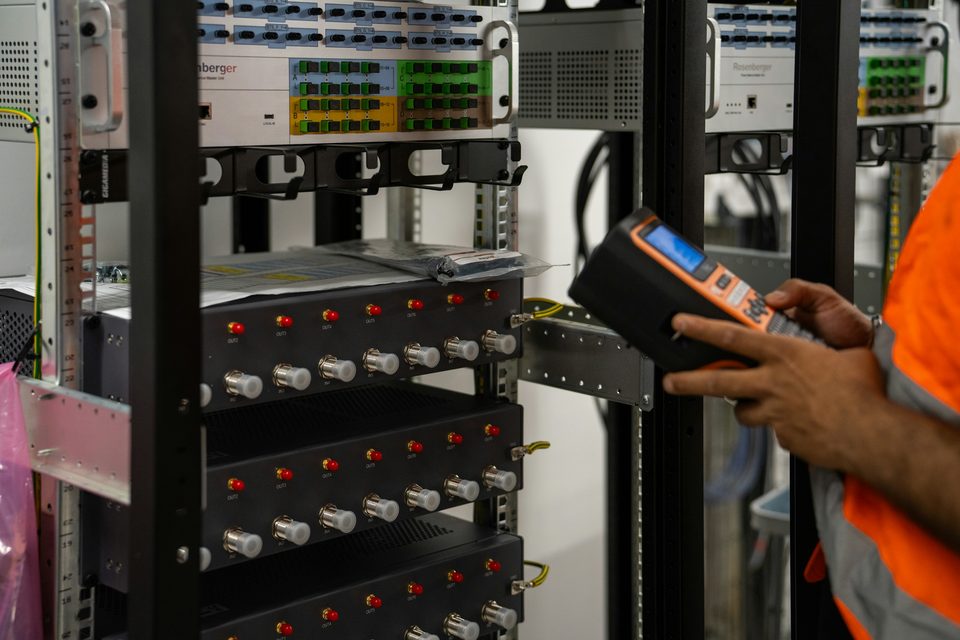 A technician in an orange high-visibility vest holds a handheld test device next to a rack of network equipment with fibre patch panels.