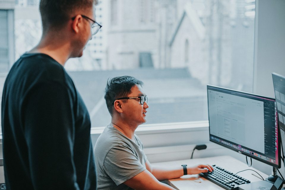A man standing by a window looks at a monitor while a seated colleague in glasses types at the keyboard and talks to him.