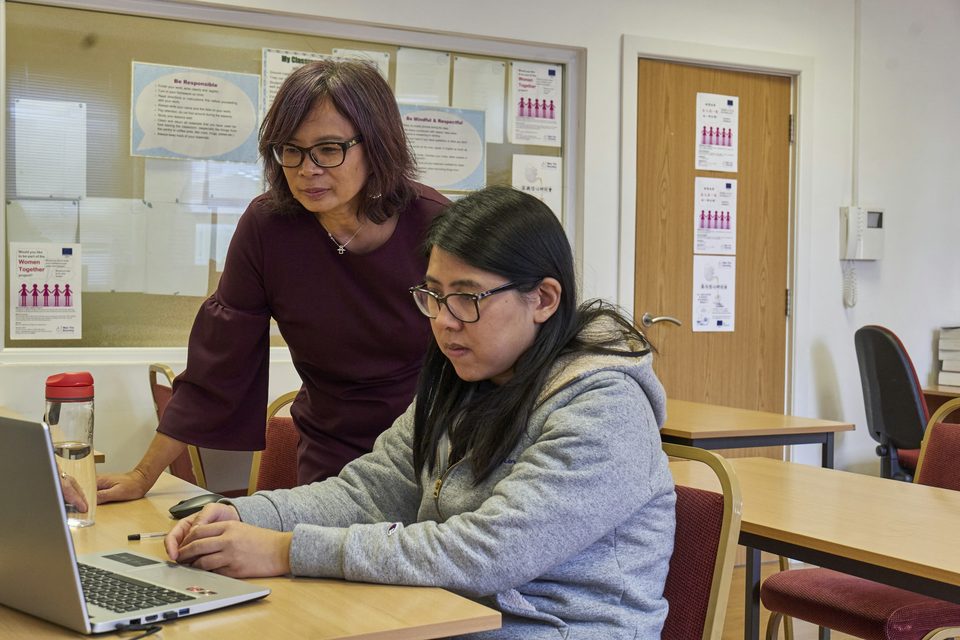 An older woman leans over to guide a younger woman working on a laptop at a desk in a classroom-style room, with a notice board behind them.